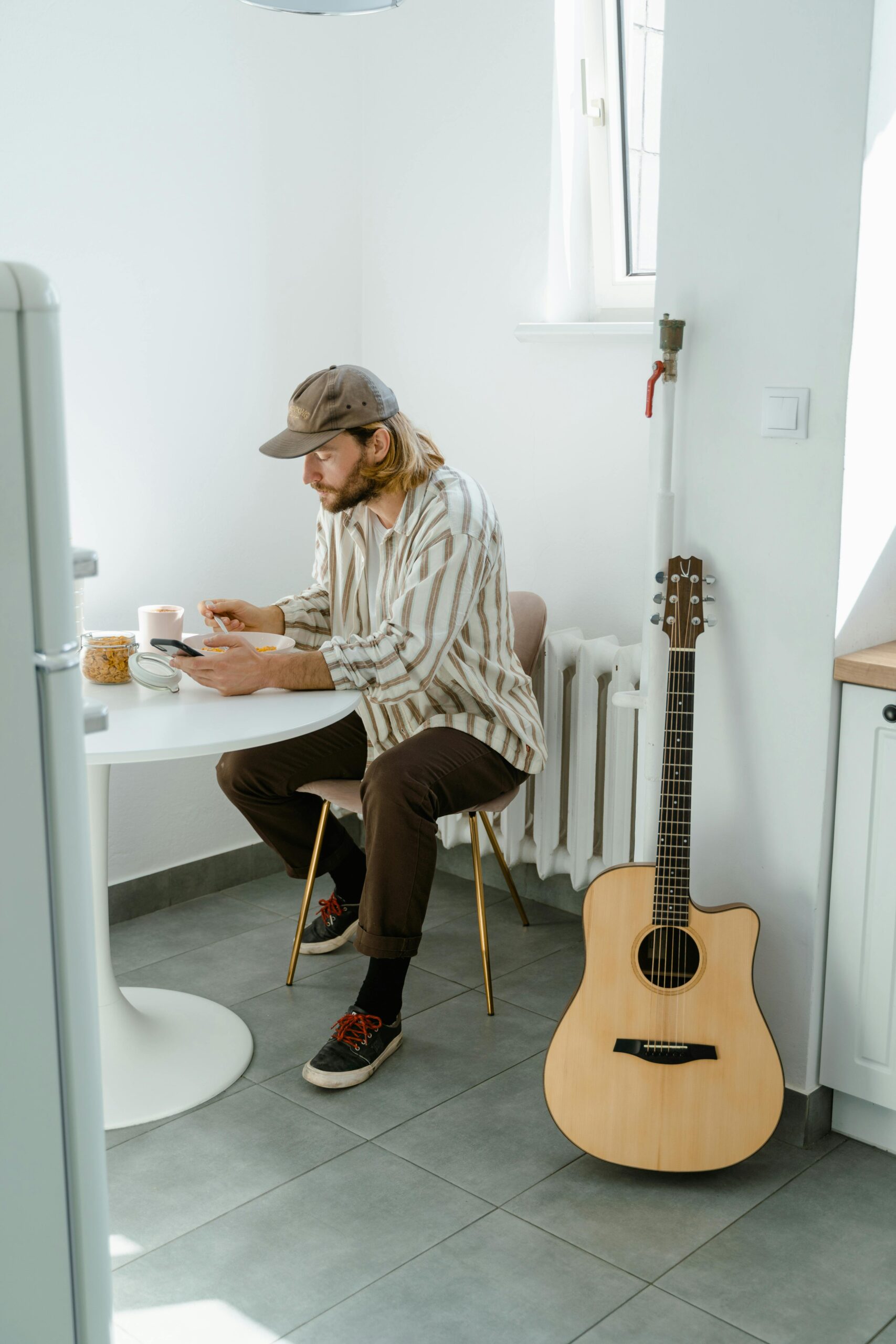 A man seated at a table in a cozy kitchen, enjoying coffee alongside an acoustic guitar.